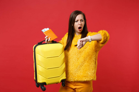 震惊的年轻女子穿着黄毛衣晚看手表，拿着旅行箱护照登机牌票孤立在红色背景上。人真诚的情感，生活方式的观念..把复制空间