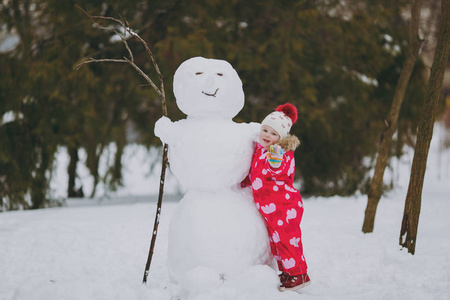 可爱的小女孩穿着冬天温暖的衣服，帽子站在雪人附近，在雪地公园或森林户外挥动手。冬季好玩，节假日休闲..恋爱关系家庭童年生活方式