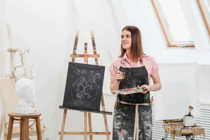 一位年轻的女画家在明亮的白色工作室里在画架上的画布上画一幅画