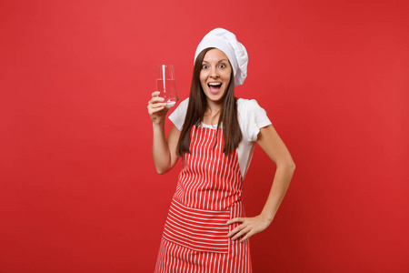 家庭主妇女厨师或面包师穿条纹围裙，白色T恤，烤厨师帽子隔离在红墙背景上。 女人拿着透明的新鲜的纯净的从玻璃中提取出来的模