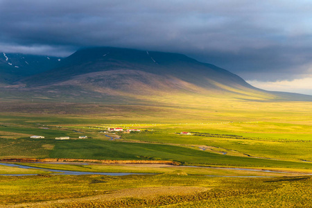 宁静的乡村景观，山脚下有农田，广阔的绿色草地，河流和乌云，在温暖的金色夏日灯光下。 欧洲冰岛农村全景图