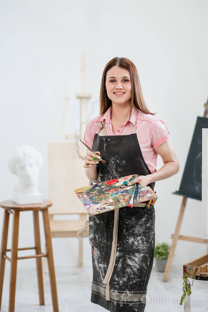 一位年轻的女画家在明亮的白色工作室里在画架上的画布上画一幅画