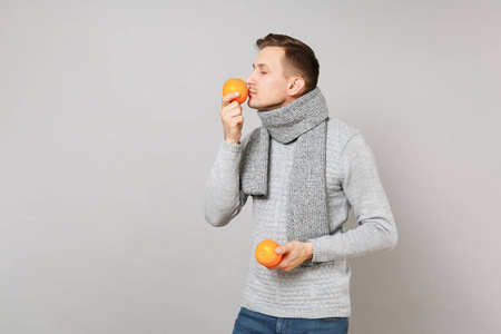 穿灰色毛衣的年轻人，戴着闭着眼睛的围巾，嗅着灰色背景上孤立的橘子。健康时尚生活方式，人真诚情感，冷季理念..把复制空间