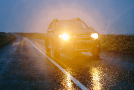 湿沥青路上的SUV汽车在黑暗的雨天驾驶。焦点之外的乡村景观，雨滴在大灯的光线下