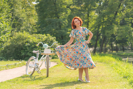 可爱的白种人女孩，红色的头发，穿着复古的连衣裙，享受着白色老式自行车的生活，在夏日的城市公园里时髦地散步。
