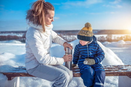 一位年轻的母亲用热汤或茶打开一个热水瓶, 和她的儿子, 一个25 的男孩坐在长凳上。在大自然中的冬日早晨休息