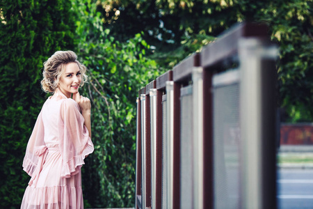 迷人的年轻女孩在一个短的粉红色礼服摆姿势附近的钢栅栏上的绿色树叶的背景