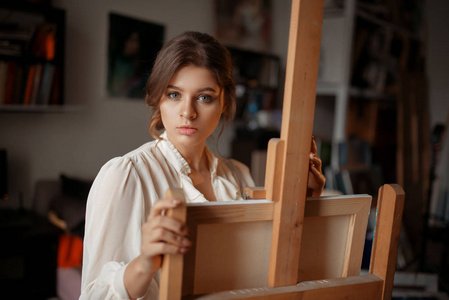 在工作室的画架上有思想的女艺术家。 创意油漆画家绘制艺术作品车间内部背景