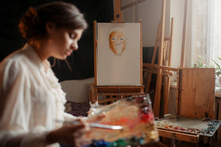 女艺术家与调色板和刷子站在画架在工作室。 创意油漆工绘画肖像车间内部