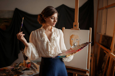 女艺术家与调色板和刷子站在画架在工作室。 创意油漆工绘画肖像车间内部