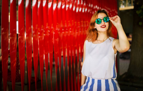 迷人的红发女人戴着太阳镜在红色背景下摆姿势。
