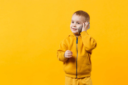 小酷童男孩，34岁，穿着黄色衣服，在手机上交谈，孤立在橙色墙背景工作室的肖像上。 儿童和小玩意的问题。 生活方式的概念。 模拟复