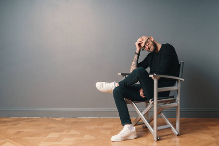 一个现代时尚的男人坐在腿上，在灰色的墙壁背景下，在工作室的一张银色椅子上摆出一个自信的姿势。 他戴着胡须眼镜，黑色便服和白色运动