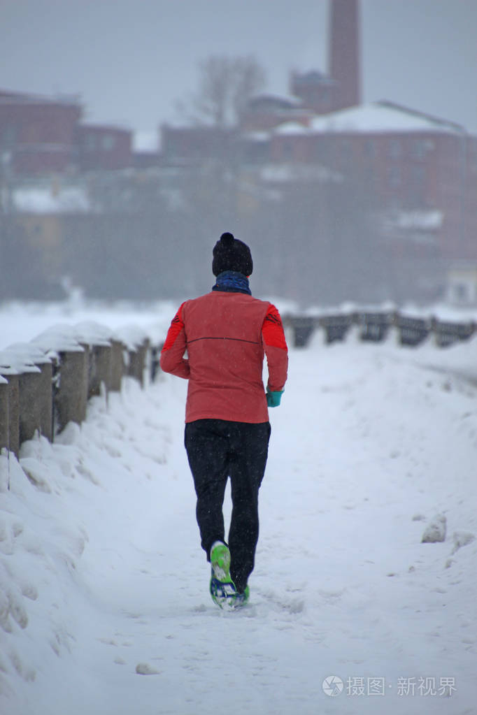 一个穿着红色夹克的人在大雪中沿着冬天的城市堤岸奔跑