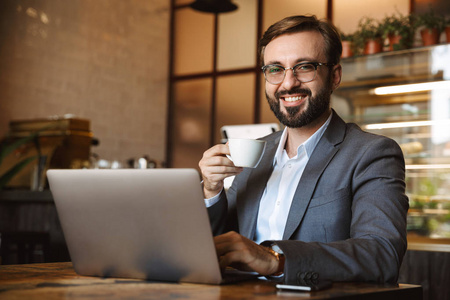 英俊的年轻商人穿着西装，在笔记本电脑上工作，坐在室内咖啡馆喝咖啡