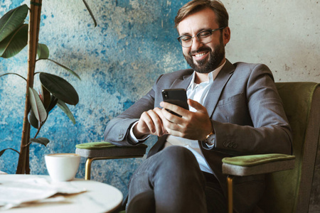 微笑的商人穿着西装拿着手机坐在咖啡馆喝咖啡