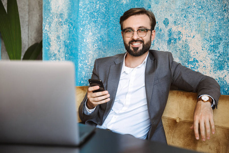 英俊的年轻商人穿着西装，在笔记本电脑上工作，坐在室内咖啡馆里，拿着手机