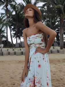 漂亮的黑发女孩，长发卷曲，头上戴着帽子，穿着夏天的连衣裙，沙滩上站着鲜花。望着平静广阔的海洋。背景上绿色的高棕榈树