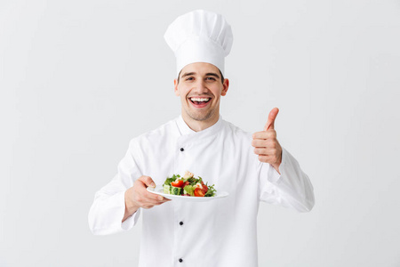 令人兴奋的男厨师穿着制服，在白色背景的盘子上展示新鲜的绿色沙拉，竖起大拇指