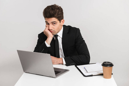 厌倦了无聊的年轻商人，穿着一套灰色背景的西装，坐在办公桌前用笔记本电脑工作