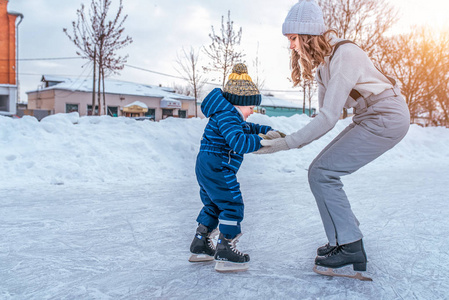一个35岁的小男孩正在滑冰。妈妈让她的儿子帮助抵抗。溜冰鞋的第一步。在城市的冬天在溜冰场上。儿童教育中的支持和支持概念