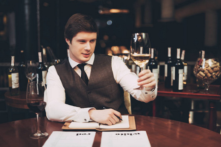 自信的侍酒师在餐厅制作葡萄酒卡片, 做笔记, 看一杯白葡萄酒