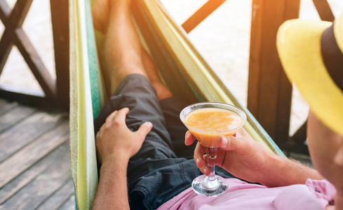 英俊的年轻人躺在海边阳光明媚的海滩上的吊床里, 手里拿着鸡尾酒。阳光明媚