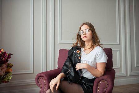 年轻时尚的模特戴着带皮夹克的眼镜摆在扶手椅上