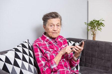 主题老人使用技术。 老灰头发的白种人女人，皱纹坐在沙发上的客厅里，用手机在手中看着触摸屏上的屏幕手指。