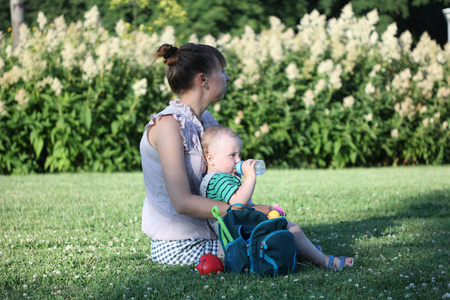 一位年轻的母亲在阳光明媚的夏日和她的孩子坐在花园里的草坪上。 那个男孩从瓶子里喝水。