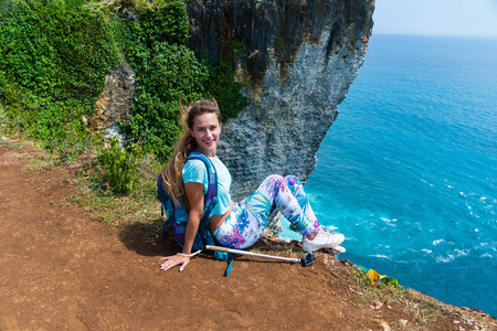 漂亮的女性旅行者背着背包坐在悬崖边上, 在镜头前微笑着。巴厘岛, 印度尼西亚