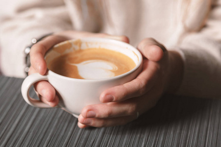 关上一杯白色的热拿铁艺术咖啡，一个年轻女孩手中有一个心形。 女人穿着温暖的冬季针织白色毛衣。 变调了。 咖啡女孩概念放松咖啡杯。