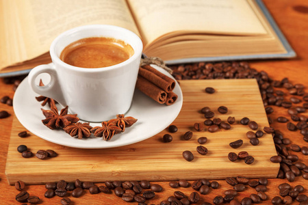 咖啡休息时间，在一张木桌上放一杯热浓咖啡，上面铺着一本敞开的旧书。一杯白色的黑咖啡，周围是少量的烤咖啡豆