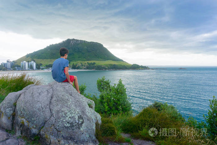 男孩坐在休闲岛上的岩石上望着大海在黄昏的灯光和背景下的乌云下