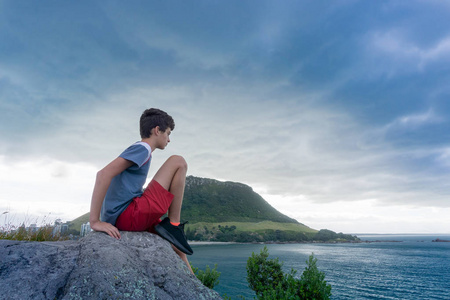 男孩坐在休闲岛上的岩石上，望着大海，在黄昏的灯光和背景下的乌云下，Maunganui山