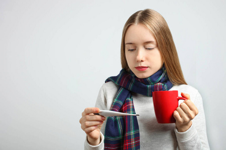 生病的白种人女孩戴着围巾，带着一杯热茶，在中性背景上用电子温度计。 概念健康热热病毒。