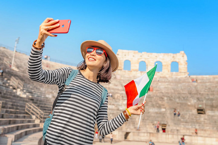 意大利古代圆形剧场前，一位快乐的女游客用意大利国旗拍自拍照片