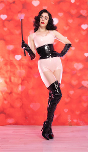 穿着粉红色乳胶橡胶服装的皮夹深色女人，穿着黑色恋物癖的紧身胸衣，膝盖以上的靴子，站着BDSM，红色背景下的作物，心。