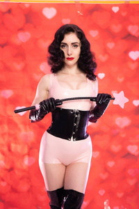 穿着粉红色乳胶橡胶服装的皮夹深色女人，穿着黑色恋物癖的紧身胸衣，膝盖以上的靴子，站着BDSM，红色背景下的作物，心。
