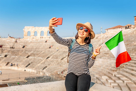 意大利古代圆形剧场前，一位快乐的女游客用意大利国旗拍自拍照片