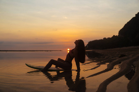 在美丽的日落背景下, 在海边的冲浪板上坐着的女孩的剪影和倒影照片