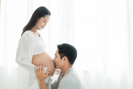 亚洲丈夫闭上眼睛亲吻妻子怀孕的肚子。 亚洲已婚夫妇和家庭观念。