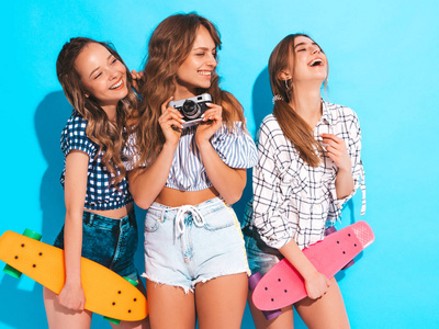 三个美丽时尚的微笑女孩和五颜六色的硬币滑板。夏天的女人靠近蓝色的墙。 拍摄复古相机