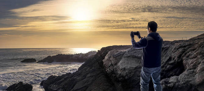 游客在日落时用手机站在加州马里布海滩海岸线上的岩石上进行移动摄影。