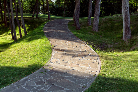 石瓦波浪式的行走小径在绿草和高大的树木之间，在一个有阳光的树林丘陵地区。