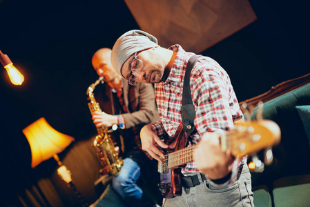 年轻的高加索人，头戴帽子，一边弹低音吉他，一边站在家里的工作室。 在背景中，萨克斯演奏家演奏他的乐器。