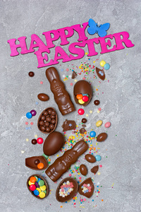 用巧克力传统鸡蛋和兔子组成的快乐复活节的粉红色文本，在灰色的混凝土背景上撒上鲜艳的悬垂和糖。