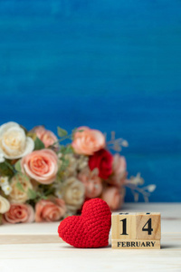 情人节的概念。 手工制作纱线红心旁边的木块日历设定在情人节日期2月14日前玫瑰花束在木桌上和蓝色背景