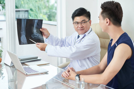 亚洲医生在医疗服装展示x射线图片的腿，年轻人坐在餐桌上