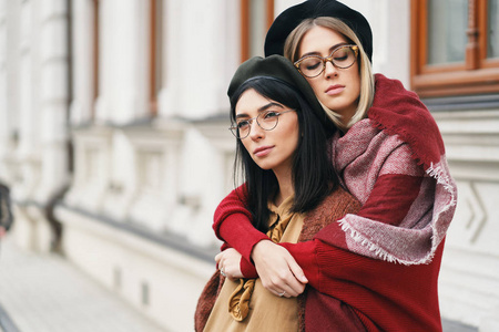 两个女朋友户外肖像。 女孩穿着休闲温暖的服装，眼镜和贝雷帽坐在城市背景复制空间上拥抱。 城市生活方式友谊和姐妹关系概念。